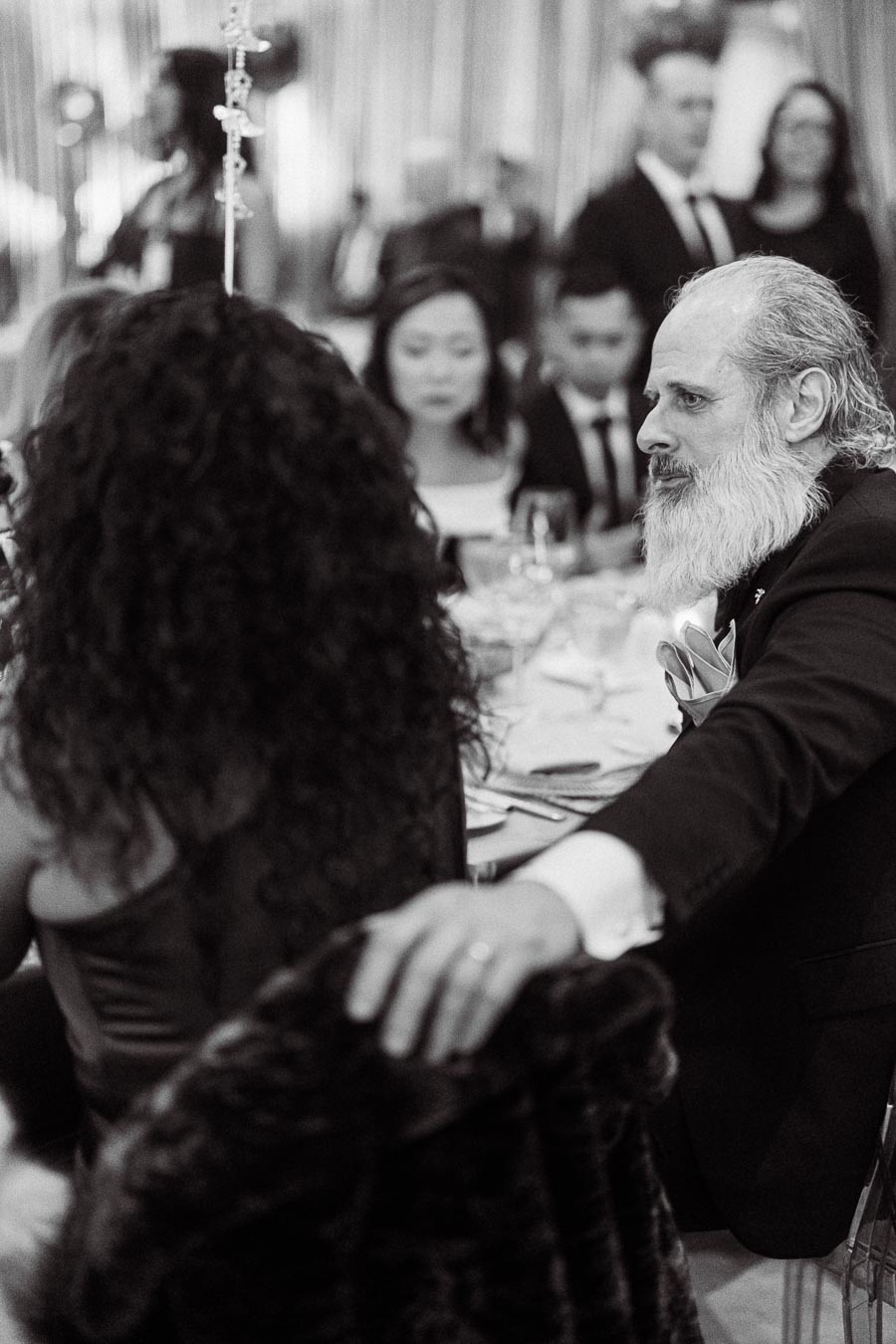 Black and white image of a bearded man in formal attire sitting at a dining table during an elegant event, surrounded by other guests.