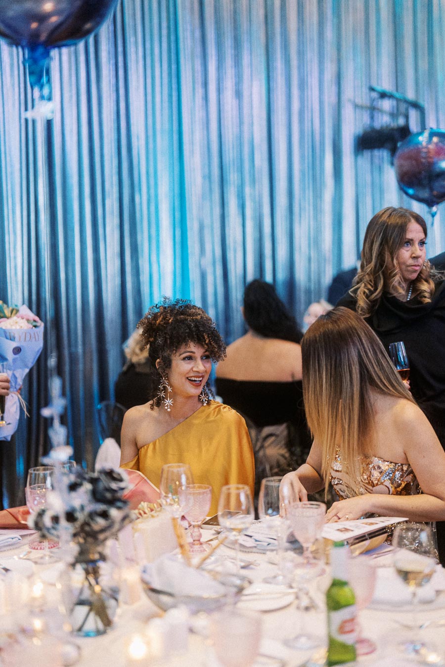 A group of elegantly dressed women sitting at a beautifully decorated table with flowers and glasses, enjoying a formal event in a venue with shimmering curtains and ambient lighting.