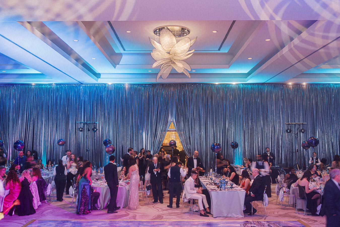 Elegant ballroom event with guests in formal attire mingling and dining under sophisticated lighting and decorative drapes.