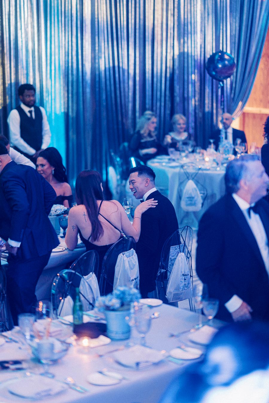 A group of elegantly dressed guests enjoying a formal dinner event, with ambient lighting and decorative backdrop, creating a sophisticated and festive atmosphere.