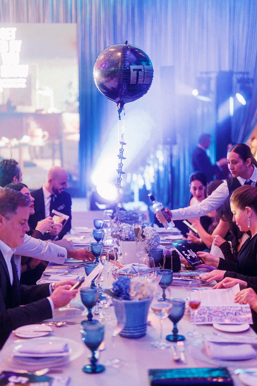 Elegant corporate event dining table setup with blue-themed decorations, assorted glassware, and a floating branded balloon centerpiece. Attendees seated around the table are engaged in conversation, enhancing the event's lively atmosphere.