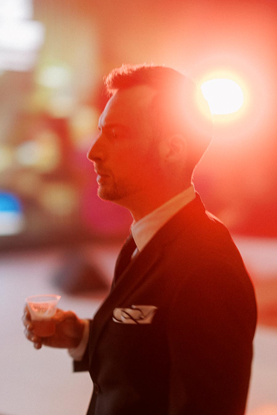 Man in a suit holding a drink, standing in a brightly lit setting with a warm, glowing background.