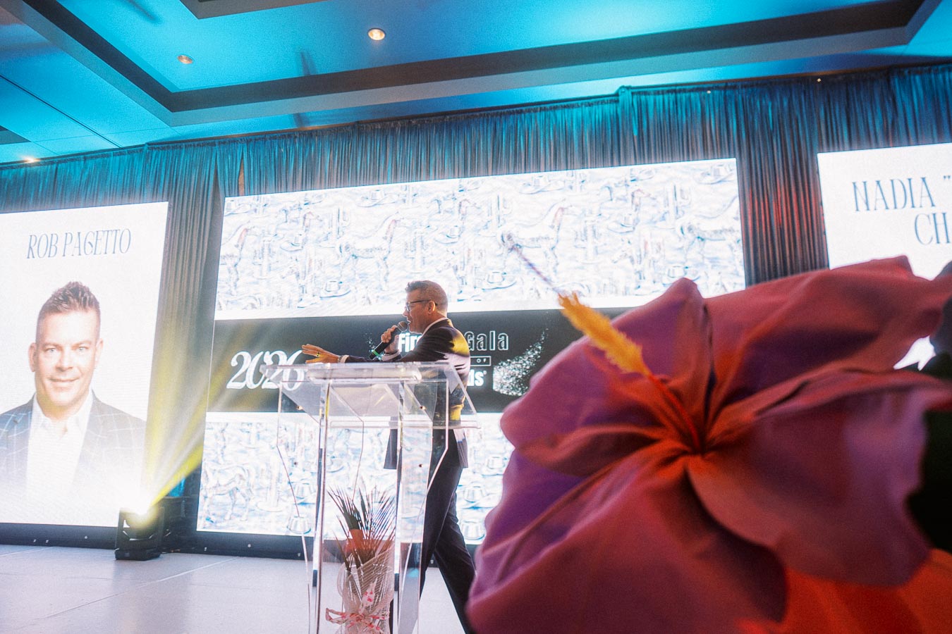 A person speaks at a podium during a professional event, with a large screen displaying images and text in the background. The room is lit with cool blue tones, and a vibrant flower is visible in the foreground.