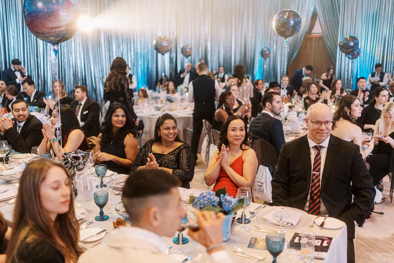 A formal event with well-dressed guests seated at decorated tables, featuring balloons and dim lighting in an elegant setting.