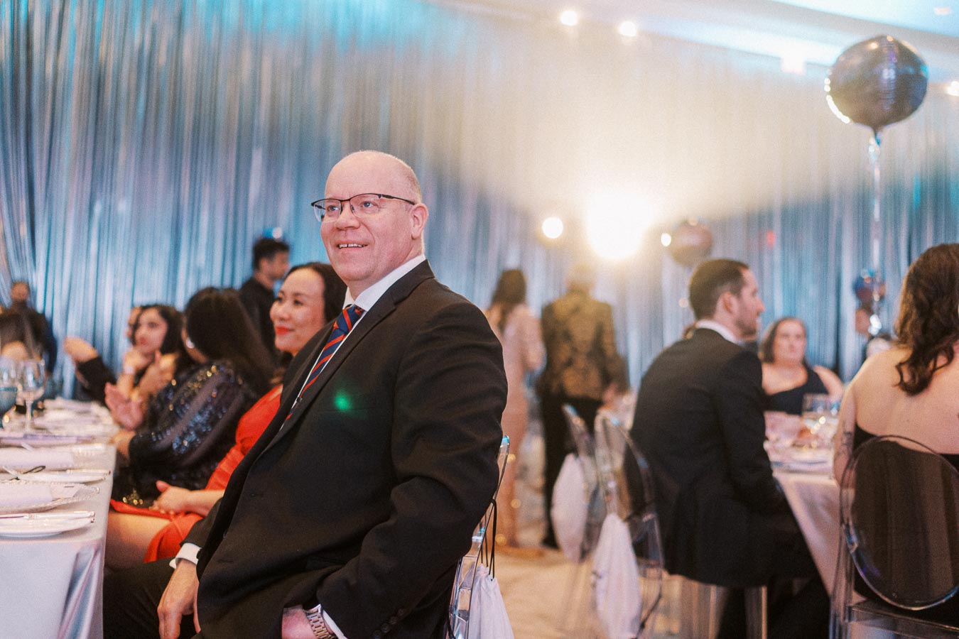 Man in formal attire at an elegant event, sitting at a decorated table with other attendees, surrounded by shimmering curtains and soft lighting.