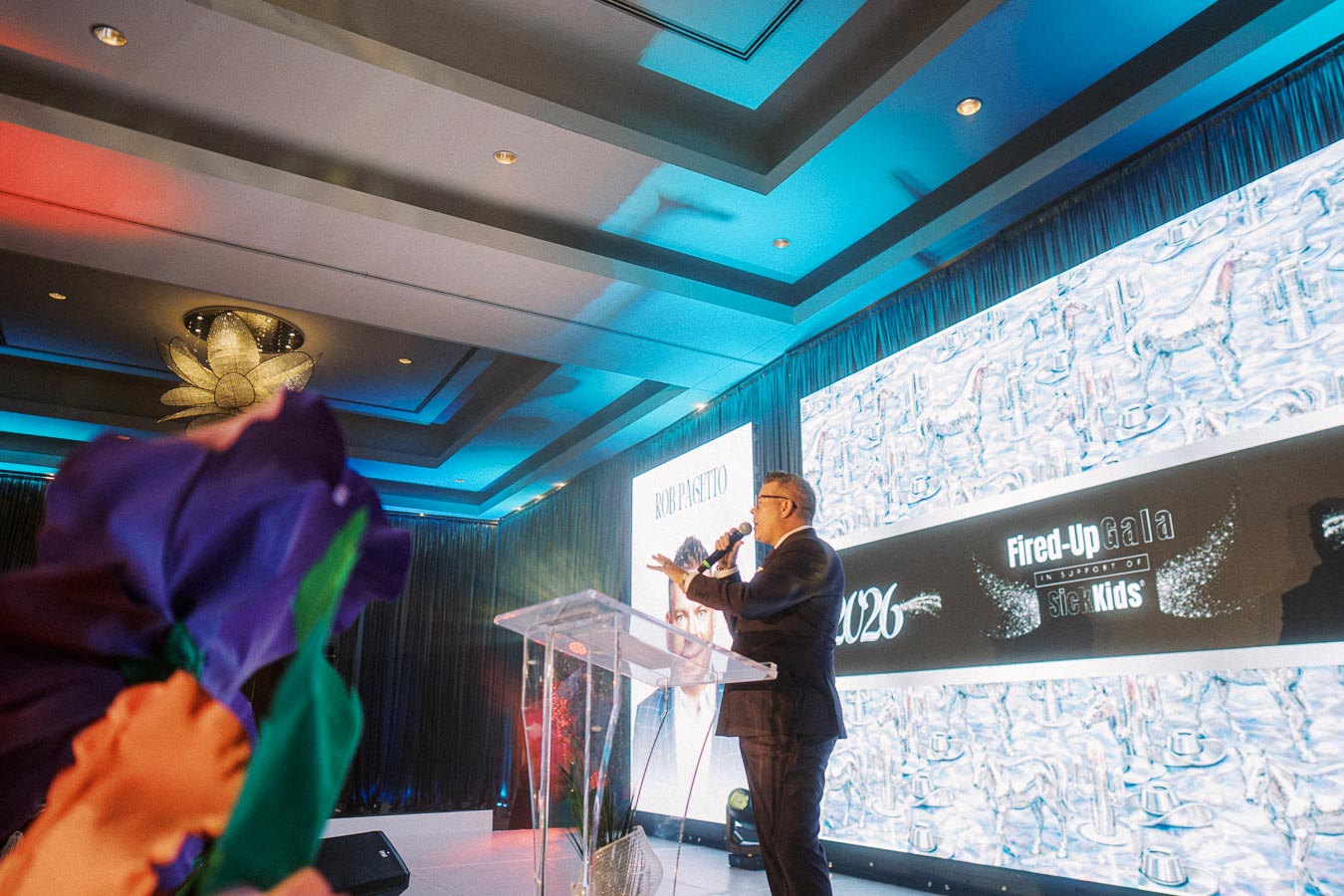 A speaker addressing the audience at the Fired-Up Gala event, holding a microphone on an elegantly lit stage with large screens displaying event graphics in a sophisticated venue.
