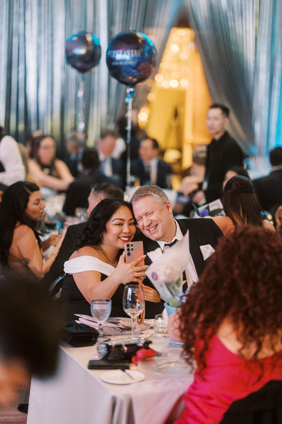 Couple taking a selfie at a formal event, surrounded by elegantly dressed guests and festive balloons.