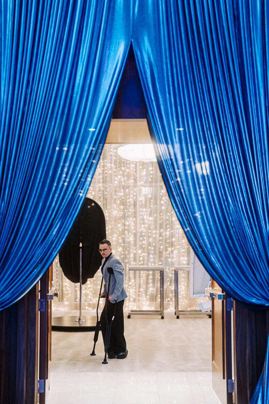 Man on crutches wearing sunglasses and a suit, framed by blue curtains with twinkling lights in the background, suggesting an elegant event ambiance.