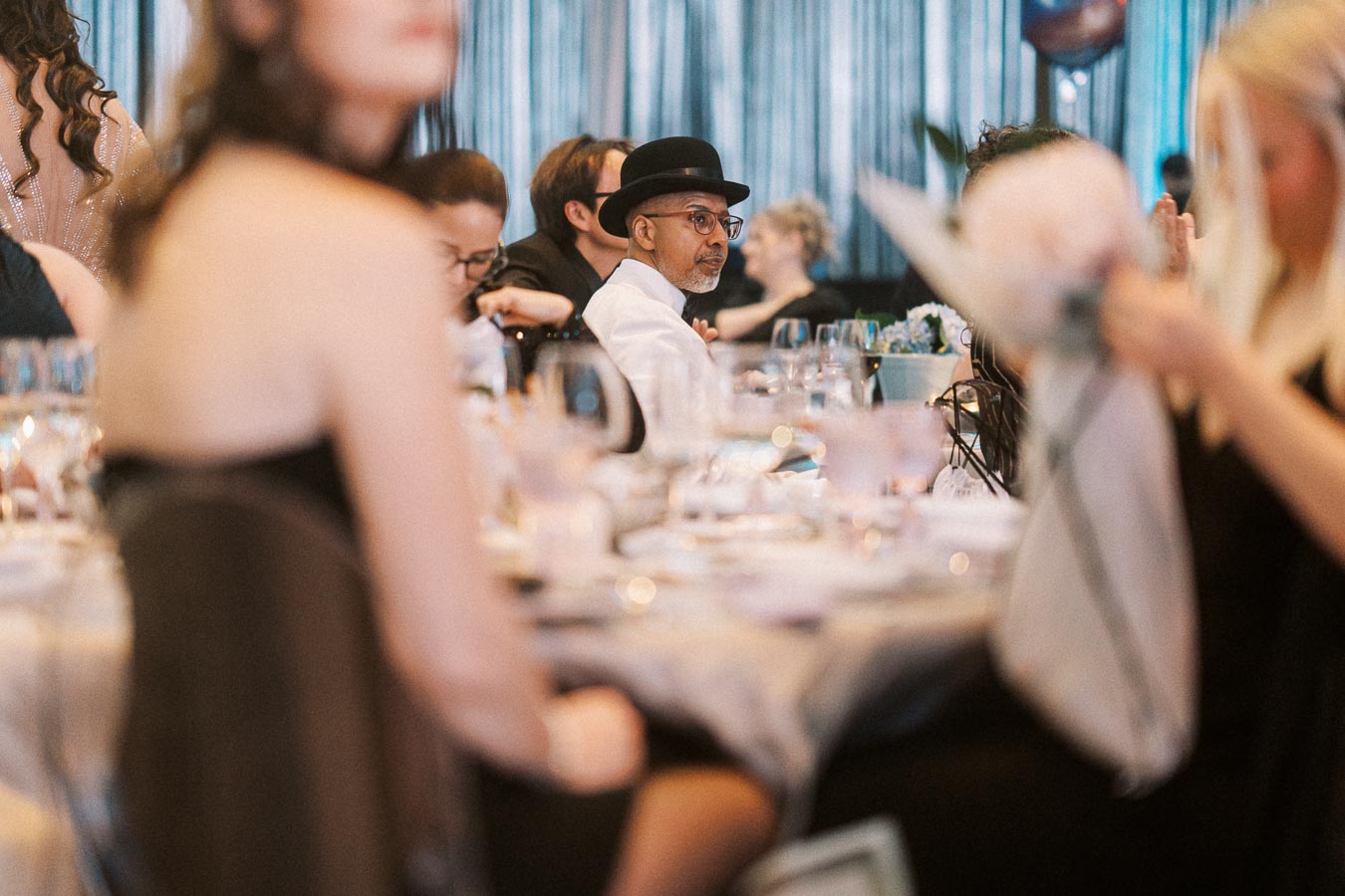 Guests seated at an elegant banquet, with a focus on a person in a black hat and glasses. The atmosphere is lively, with dining tables and a formal setting.