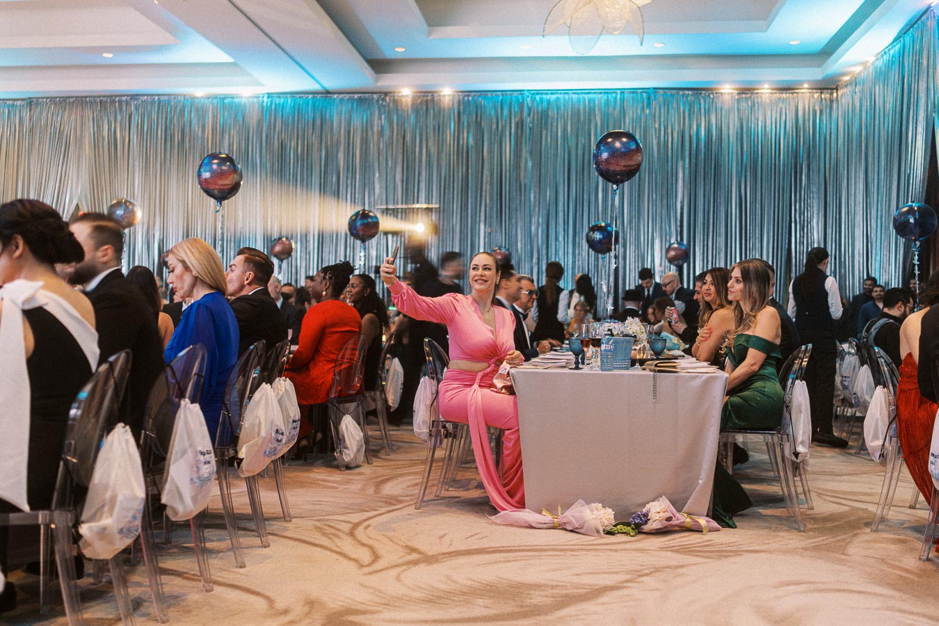 Elegant event scene with guests seated at a formal dinner. A woman in a pink dress is taking a selfie, surrounded by elegantly dressed attendees under blue lighting and decorative balloons, creating a festive atmosphere.