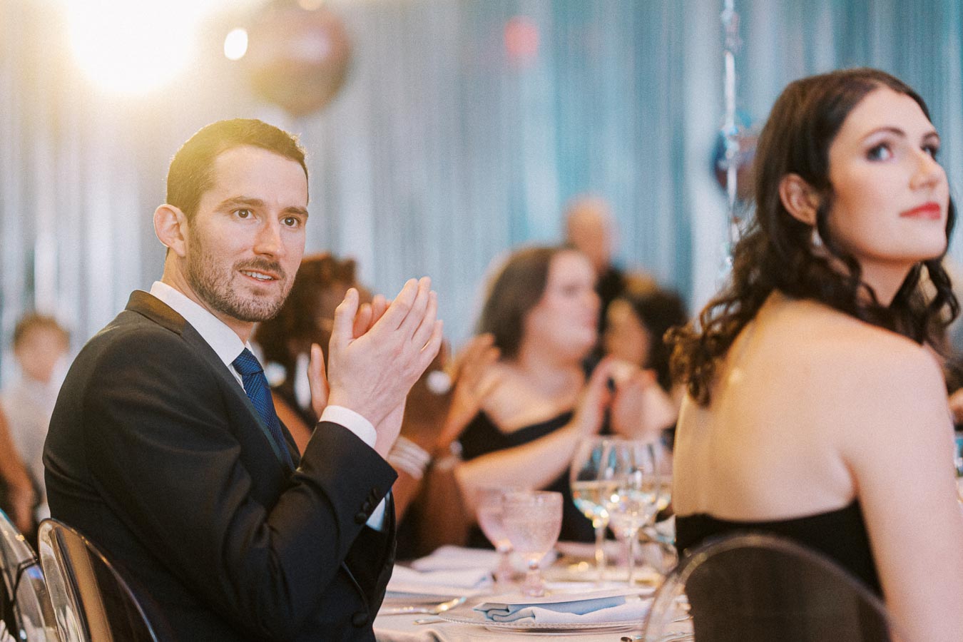 Guests dressed elegantly at a formal event, sitting at a table with glassware, as a man in a suit applauds with a bright light in the background.