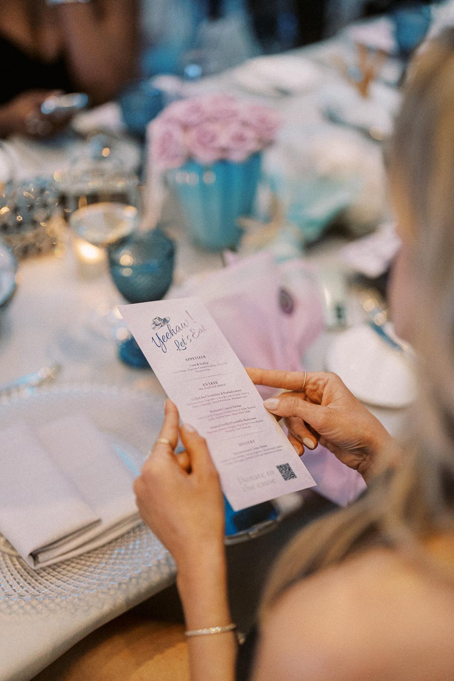 A person holding a printed menu that reads Yeehaw! Let's Eat at an elegantly set table with blue vases, pink flowers, and fine dining utensils.