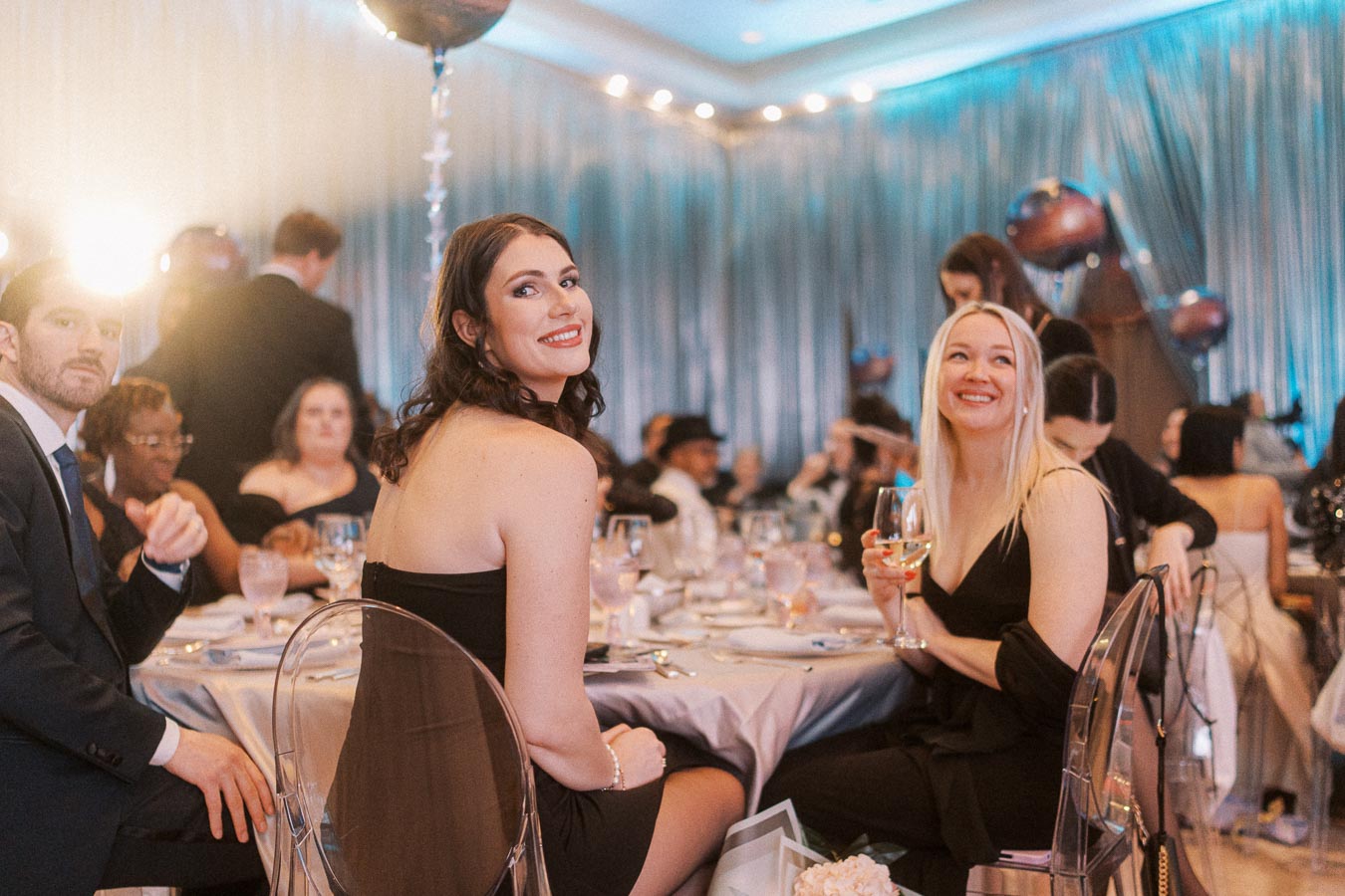 Guests enjoying an elegant evening event, dressed in formal attire, while seated at a beautifully decorated table with balloons and ambient lighting.