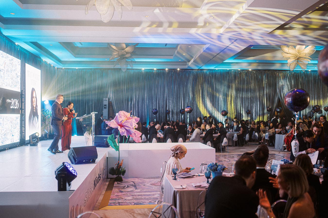 Grand ballroom event with people seated at tables, presenters on stage in front of a large screen displaying graphics, elegant lighting, and decorative elements.