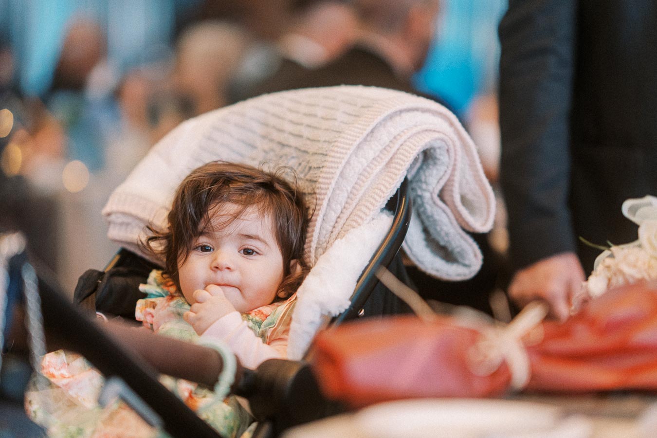 A cute toddler sits comfortably in a stroller at a bustling event, bundled with a cozy blanket and wearing a colorful outfit.