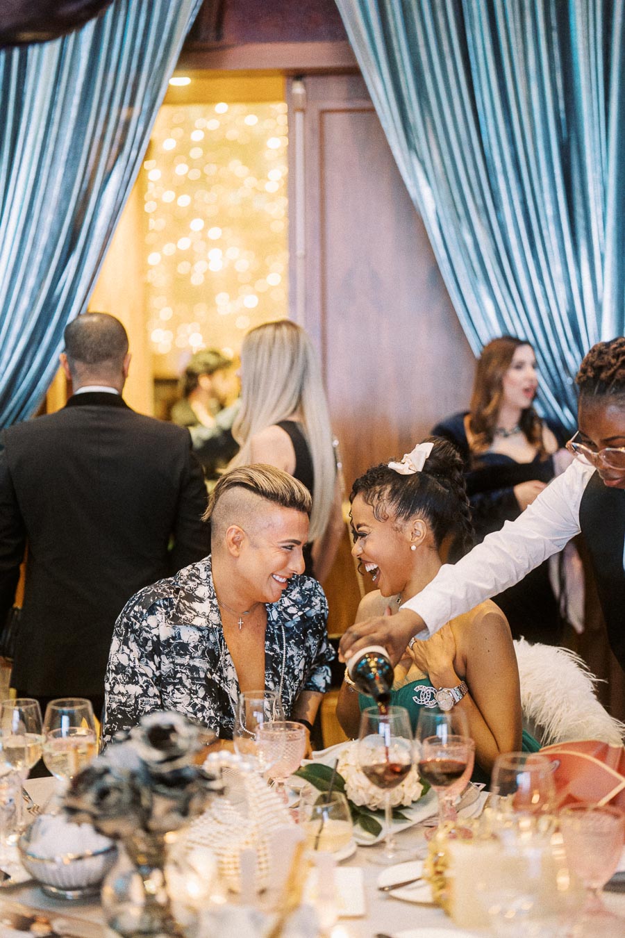 Elegant dinner party setting with guests enjoying conversation and laughter, surrounded by decorative tableware and ambient lighting. A server pours wine into a glass as part of the celebration.