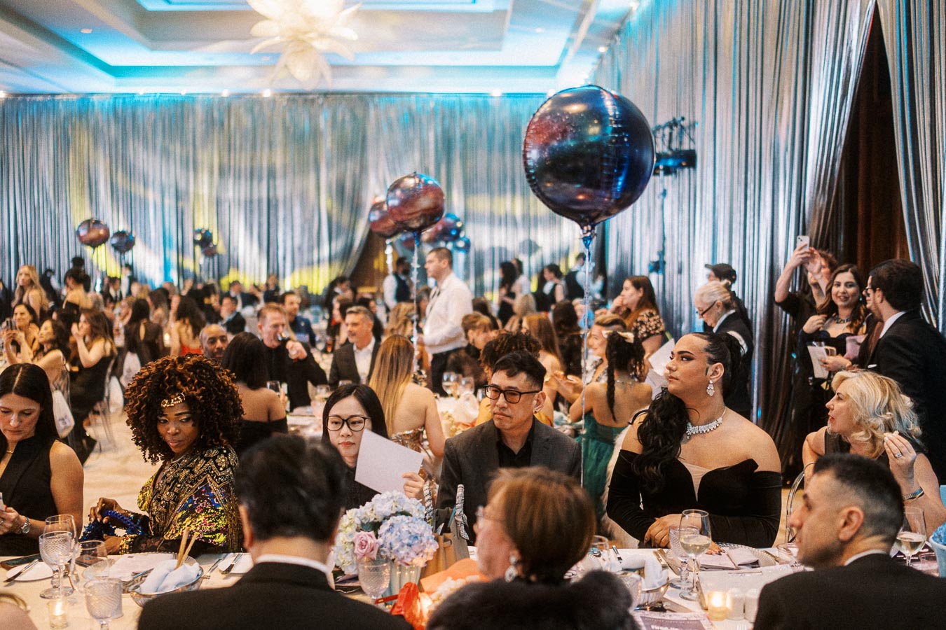Elegant indoor gala with guests seated at round tables, adorned with blue and black balloons. The room features ambient lighting and decorative drapes, creating a sophisticated atmosphere.