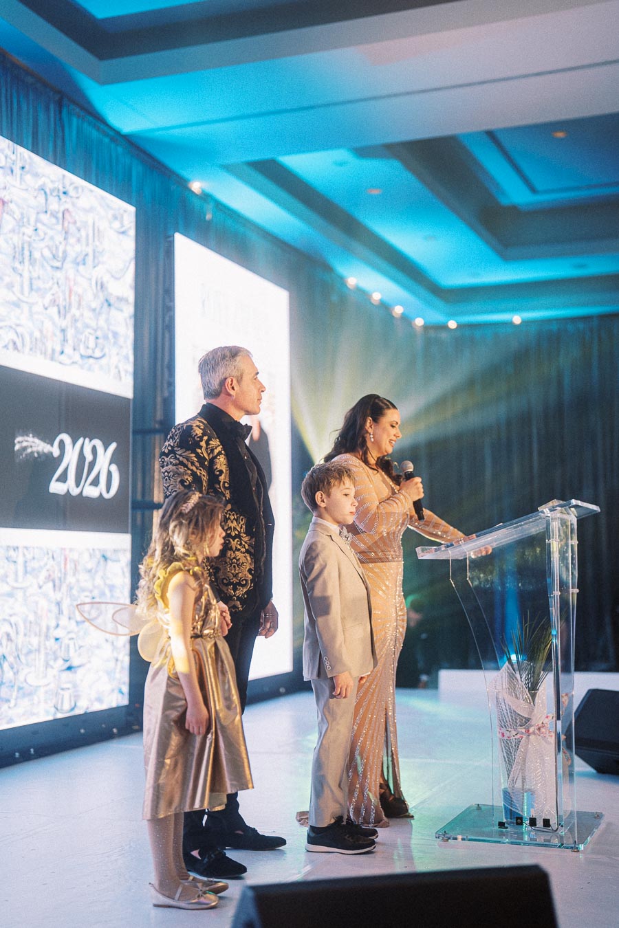 A family dressed in elegant evening wear stands on stage during a formal event, with a woman speaking at a podium and a bright 2026 displayed on a screen in the background.