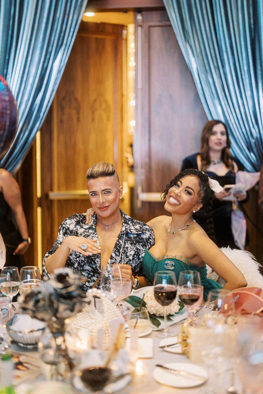 Two people smiling at an elegant event, seated at a table decorated with wine glasses and floral centerpieces, with turquoise drapes in the background.