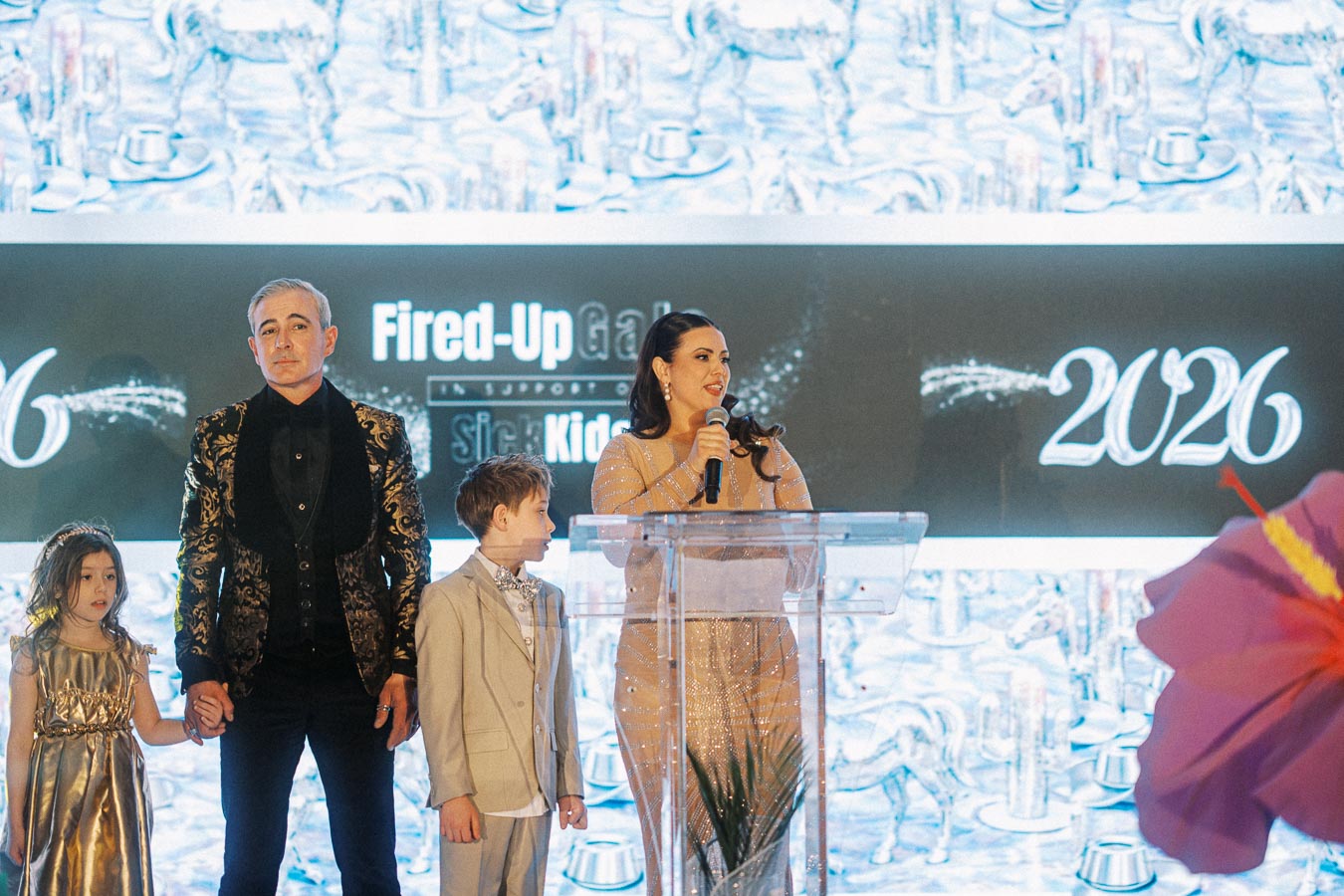 A family stands on stage at a formal event, under the backdrop of Fired-Up Gala 2026. The woman is speaking into a microphone at a clear podium, while the man and two children stand beside her, dressed in elegant attire.