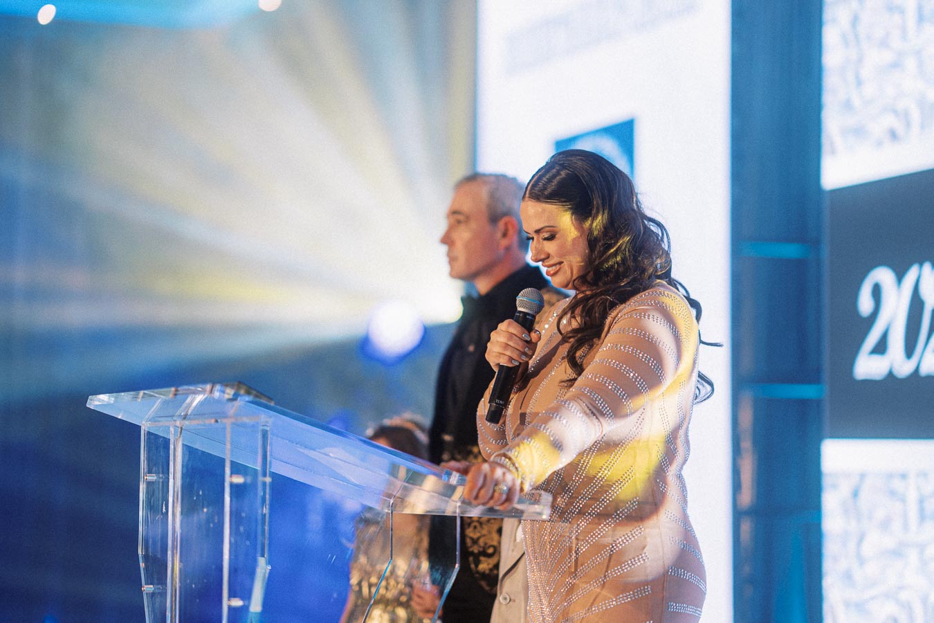 A woman smiling while speaking into a microphone at a podium during a formal event. A man stands beside her, with decorative lighting in the background.