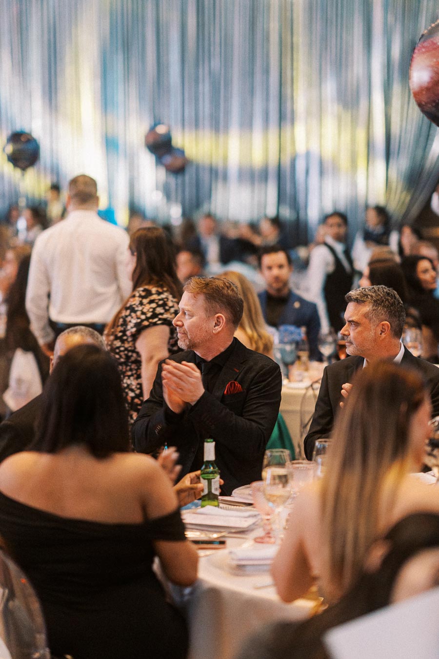 People sitting at a formal dinner event, clapping and engaged in conversation, with a decorated backdrop and festive atmosphere.