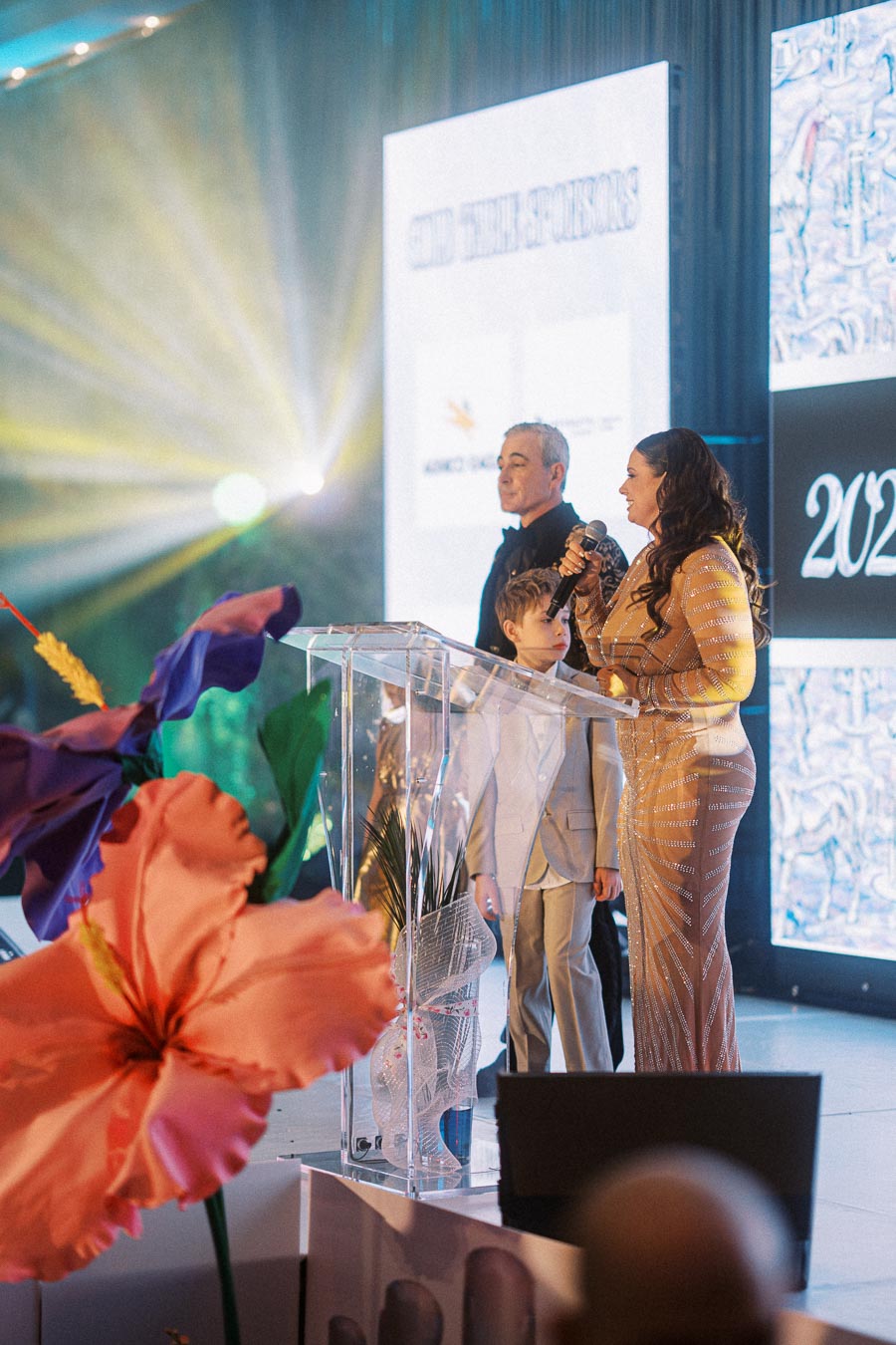 A woman in an elegant dress speaks into a microphone at a brightly lit event stage alongside a man and child, with colorful floral decorations in the foreground and event branding on digital screens behind them.