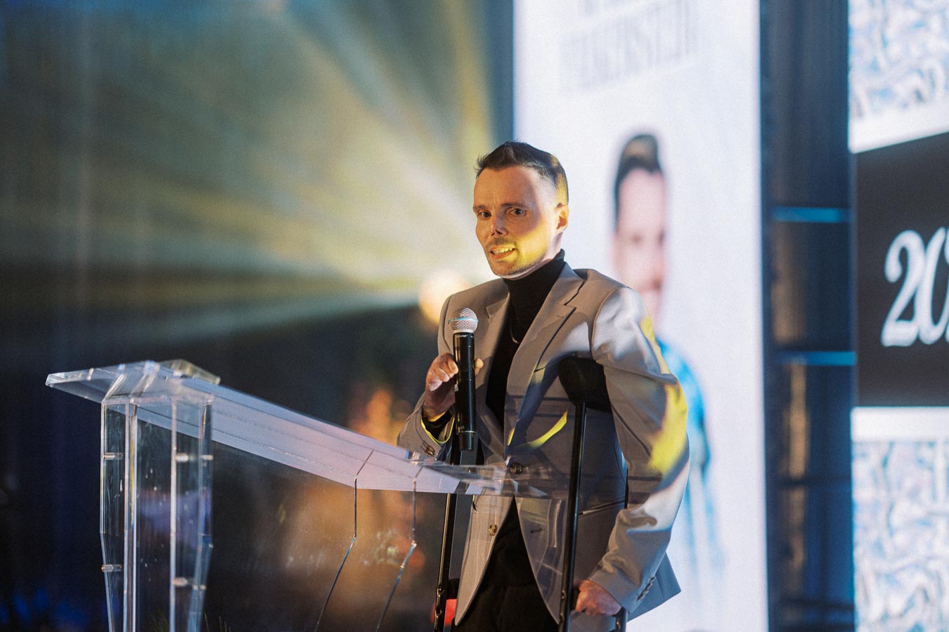 A person in a suit speaks at a podium with a microphone on stage, featuring dramatic lighting and a large screen backdrop with an image and the number 2023.