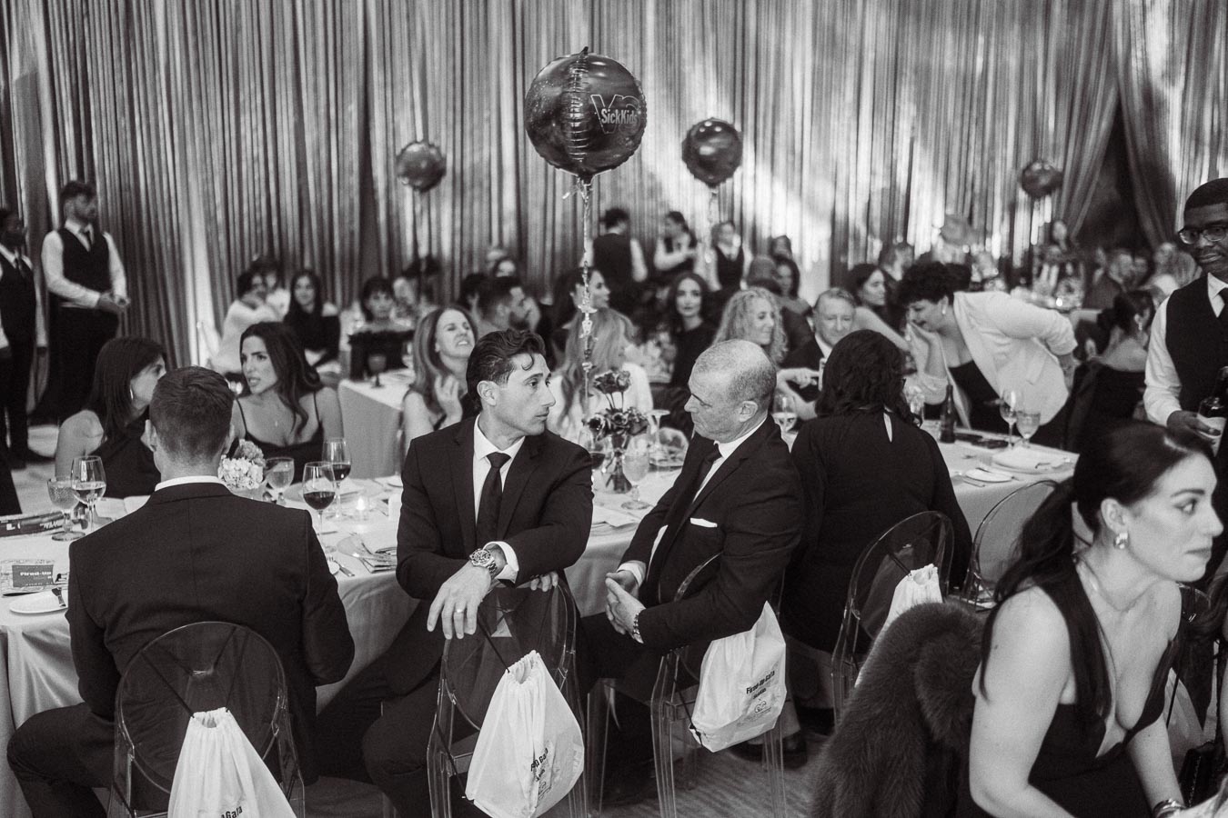 Black and white photo of a formal gala or banquet setting, featuring elegantly dressed attendees seated at tables, engaging in conversation. Balloons and decorative curtains in the background create an upscale ambiance.