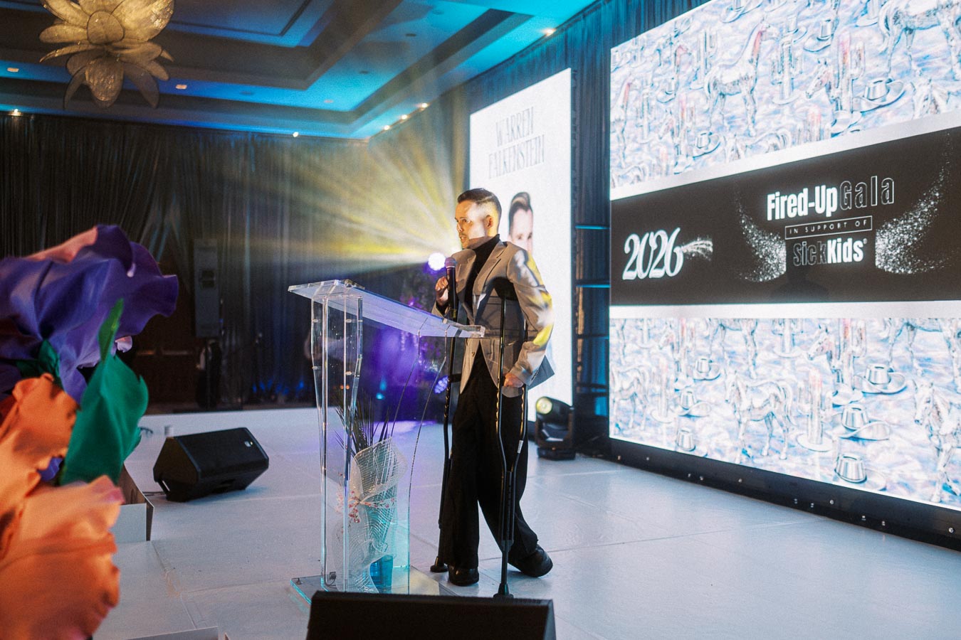 A person in formal attire speaks at a clear podium during the Fired-Up Gala event for SickKids, with illuminated screens displaying 2026 and floral decorations in the foreground, set in an elegantly lit venue.