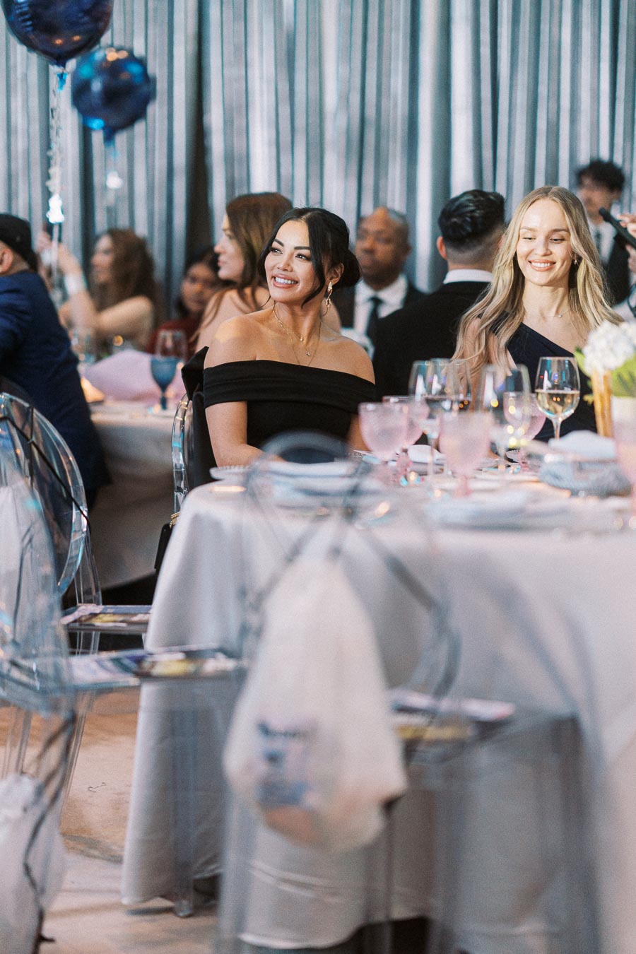 Smiling guests seated at an elegantly decorated table during a formal event, featuring clear chairs and a blue-themed ambiance.