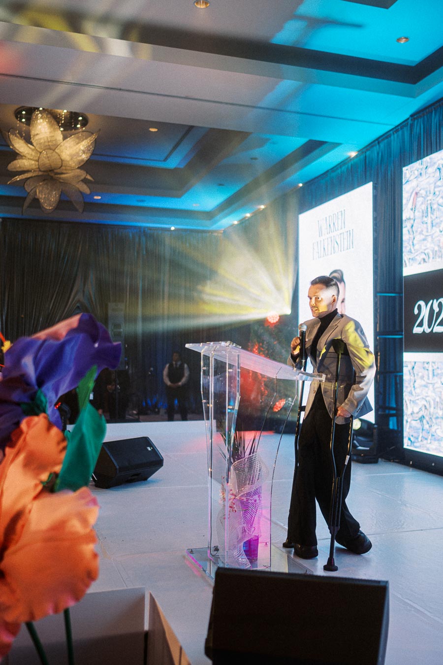 A person giving a speech on stage at a formal event, standing behind a transparent podium. The venue features vibrant lighting, with large floral decorations in the foreground and an illuminated backdrop displaying 2023.