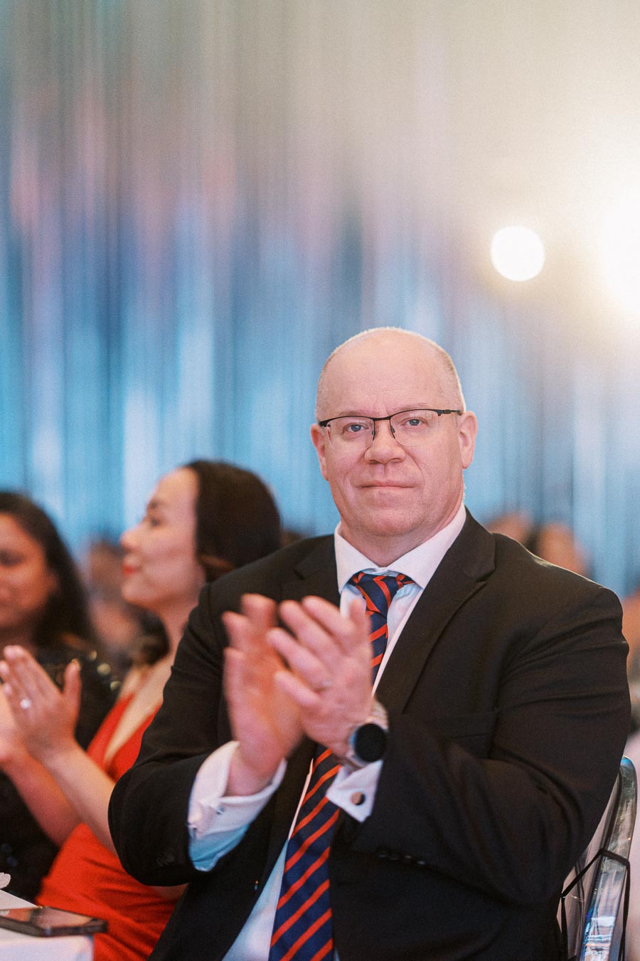 A man in a suit clapping during a formal event, with blurred attendees in the background, elegantly capturing the celebratory ambiance.