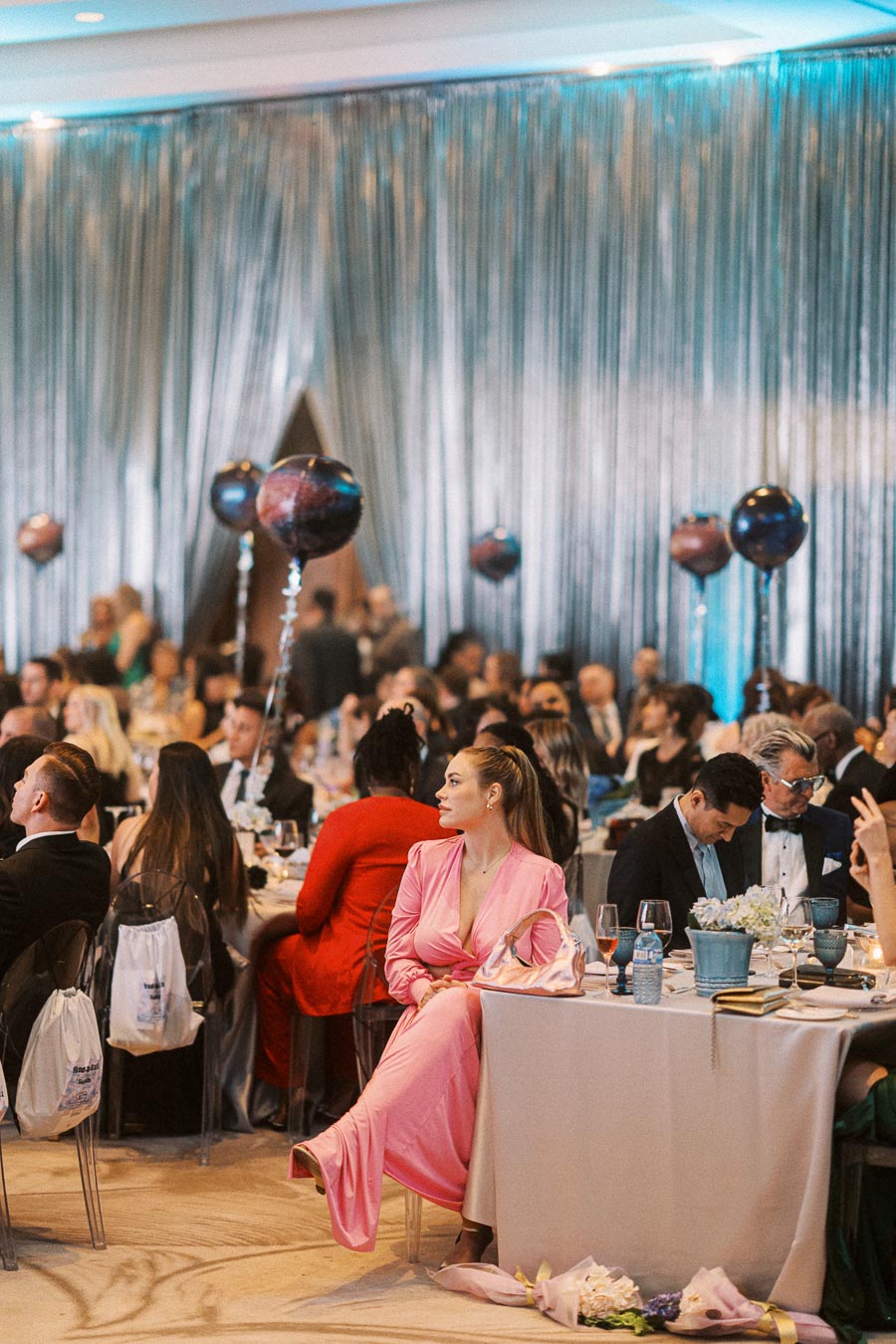 Elegant gala event with guests seated at decorated tables, featuring shimmering drapery and festive balloons, highlighting an attendee in a vibrant pink dress with classy accessories in a sophisticated setting.