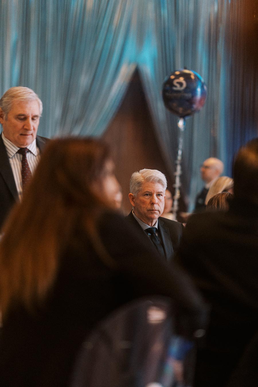 A distinguished man in a suit attending a formal event, surrounded by other guests in an elegantly decorated room with blue drapery and a themed balloon in the background.