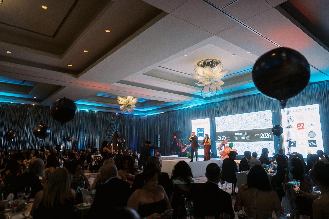 Elegant event in a luxurious banquet hall, featuring a stage with speakers and a large screen displaying various logos and graphics; attendees in formal attire are seated at tables with decorative lighting.