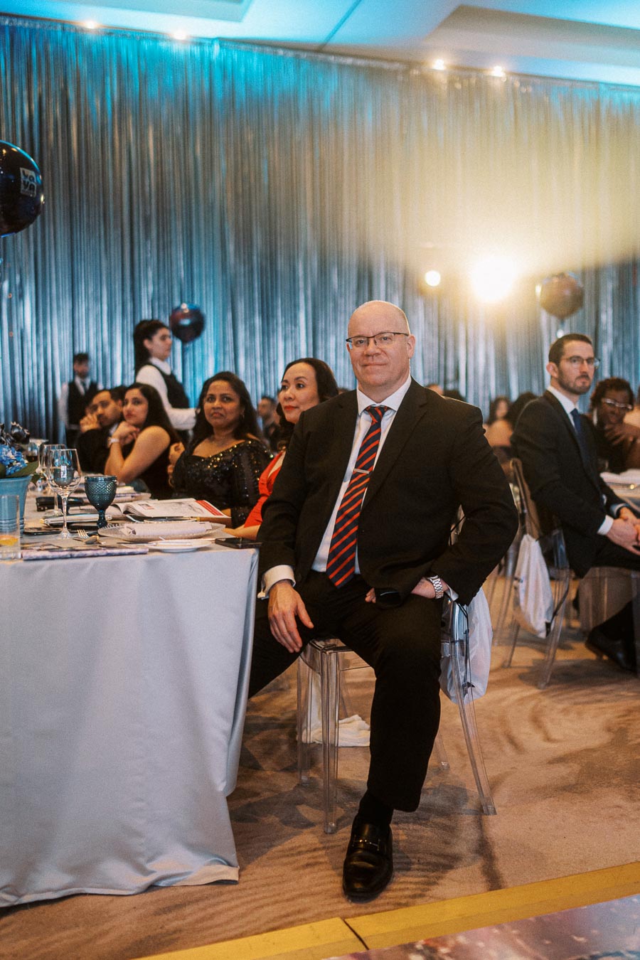 A well-dressed attendee at a formal event, seated at a decorated table in an elegantly lit banquet hall, with other guests and waitstaff in the background.
