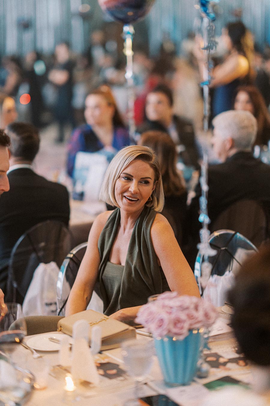 Elegant woman in sleeveless dress smiling at a formal event, surrounded by guests seated at a table adorned with a floral centerpiece.