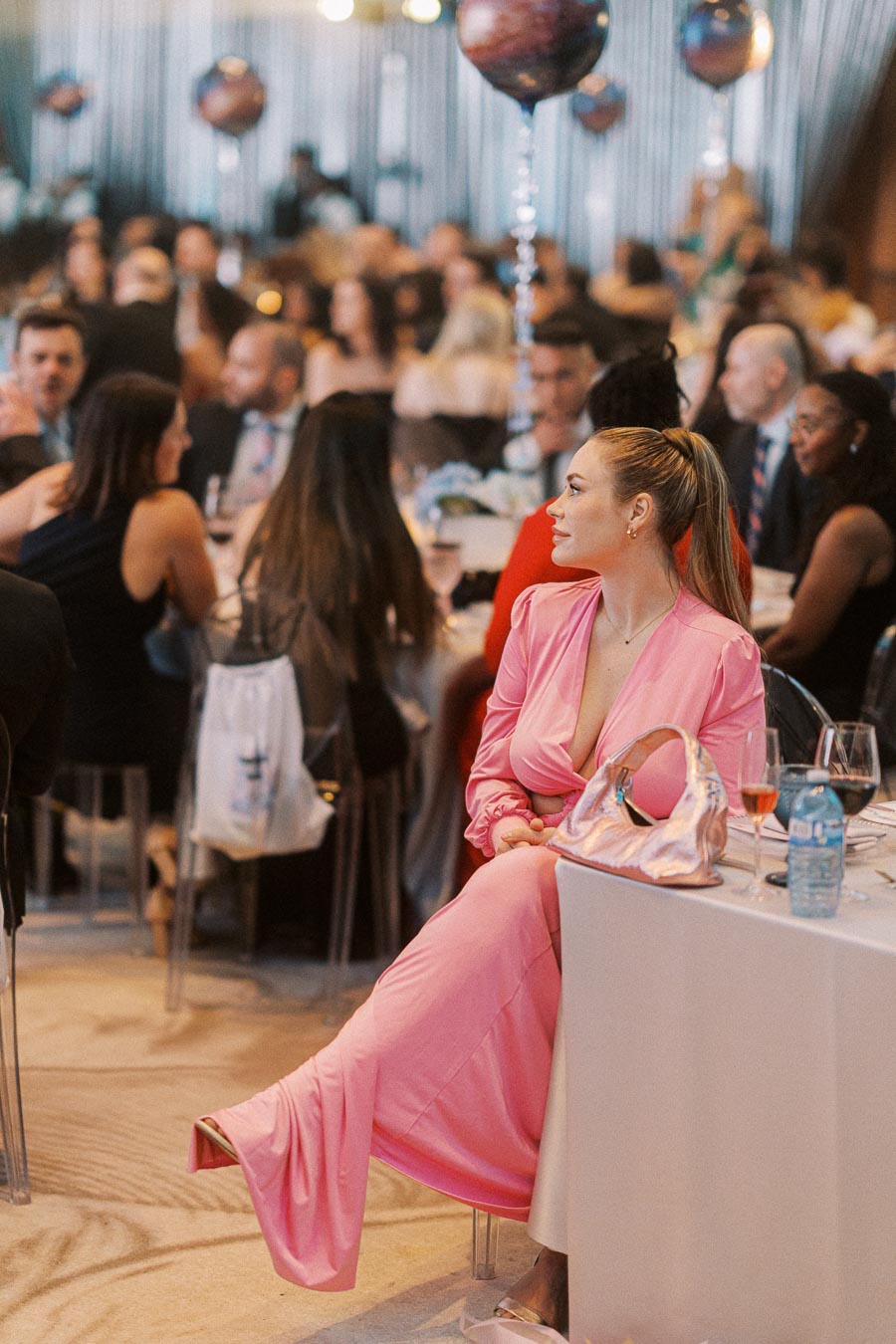 Elegant woman in a pink dress sitting at a formal event with people socializing and blurred balloons in the background.