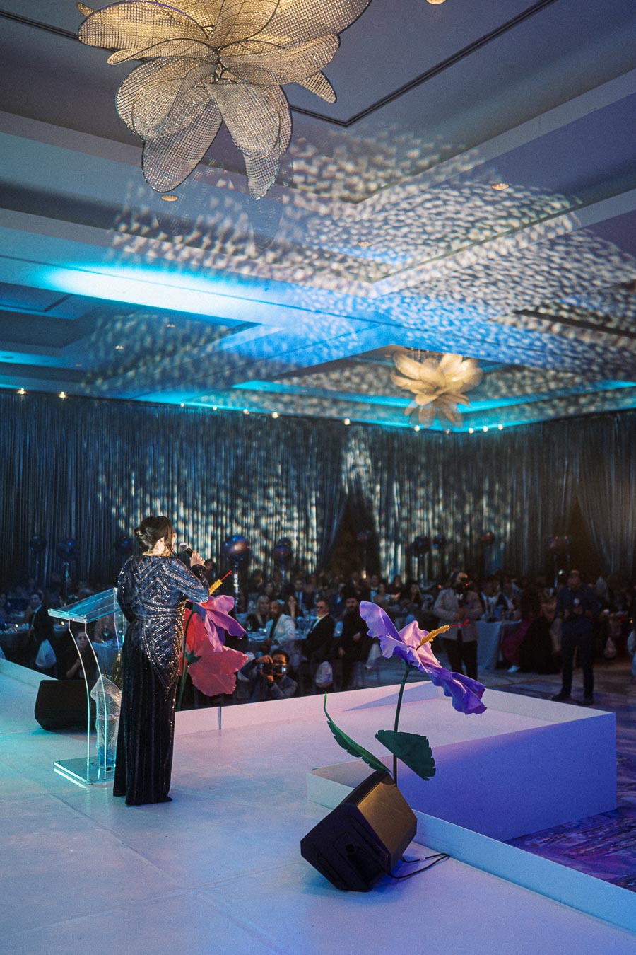 A speaker in a shimmering gown addresses an audience at a formal event, decorated with large floral sculptures and intricate ceiling lights, creating an elegant and festive atmosphere.