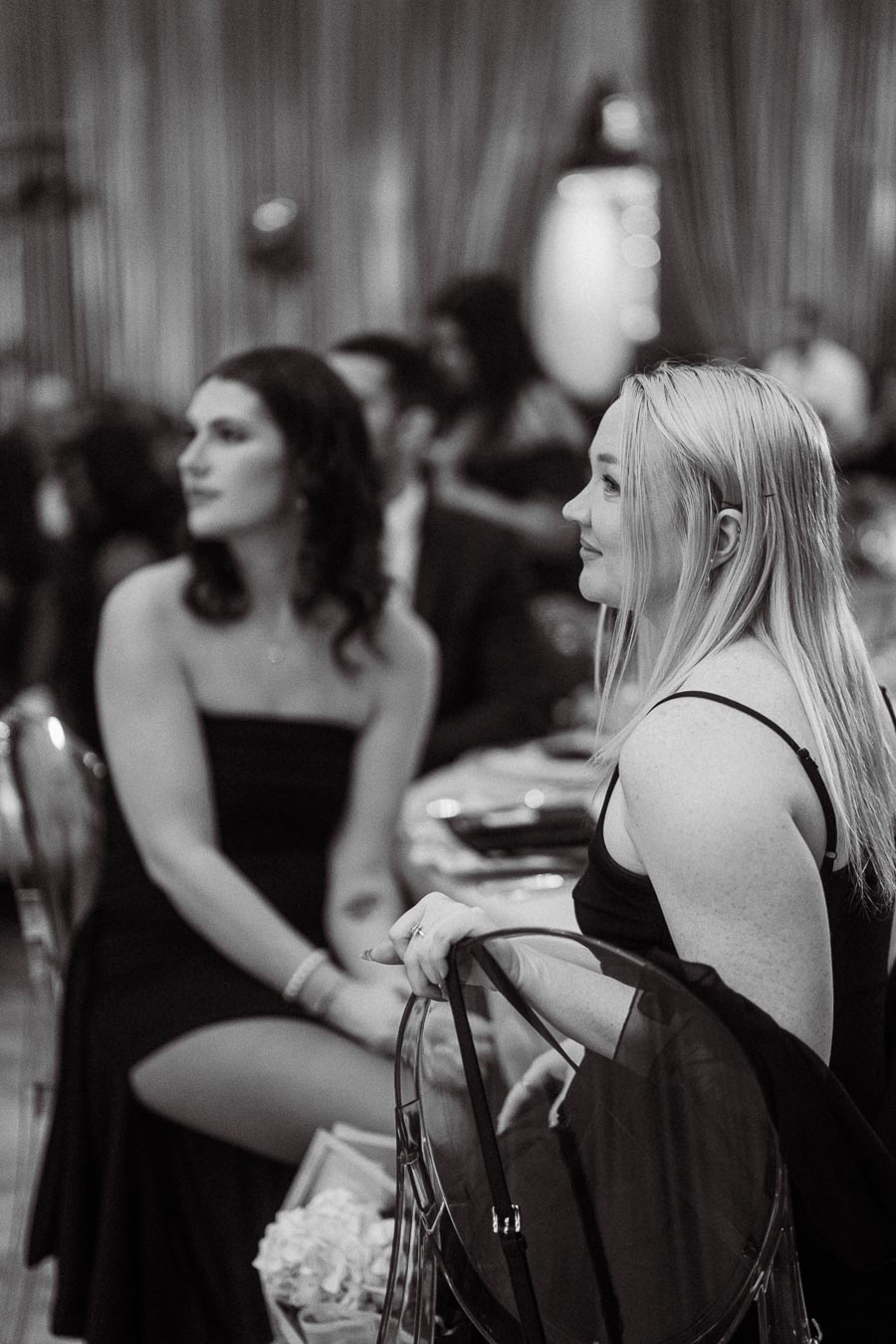 Black and white image of two elegantly dressed women seated at a formal event, attentively observing what's happening in front of them.