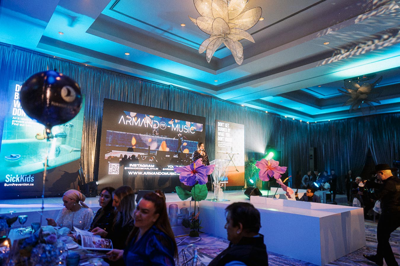 A vibrant event setting featuring a decorated stage with a presenter speaking. A display screen shows Armand Music with social media information. The room is bathed in blue and purple lighting, with attendees seated at tables and large decorative flowers in the foreground. The atmosphere is lively, reflecting a musical or celebratory occasion.