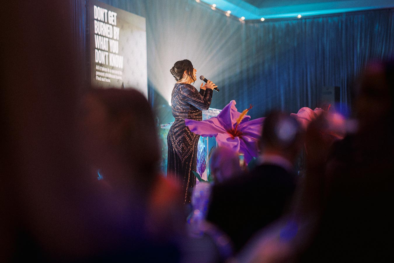 Woman in elegant dress speaking with a microphone at a formal event, backed by a large screen with text, surrounded by an audience and decorative flowers.