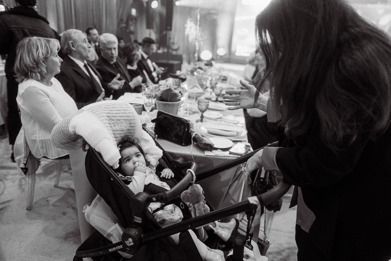 Black and white image of a formal gathering, featuring a seated baby in a stroller with adults conversing around a dining table, creating a warm family atmosphere.
