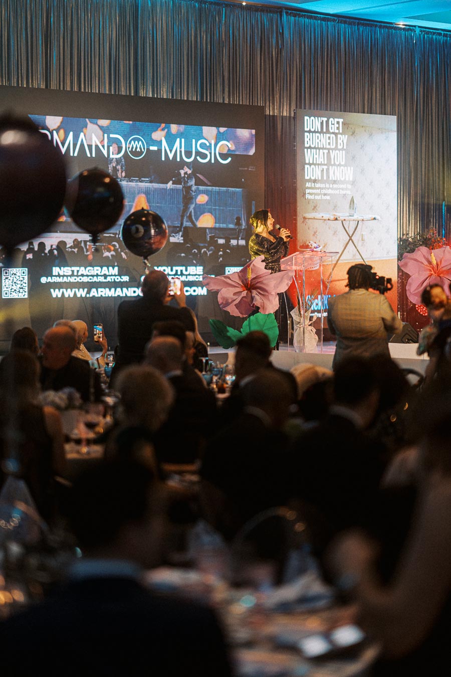 A lively event with a musician performing on stage, surrounded by an attentive audience. Large screen displays promotional material, including social media handles and website information. The decor includes vibrant flower arrangements, and a photographer and guests capture the moment.