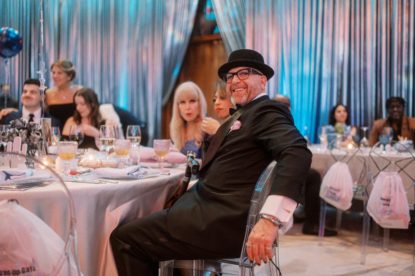 Group of well-dressed guests enjoying a lively gala event, seated at elegantly decorated tables with drinks and ambient blue lighting.