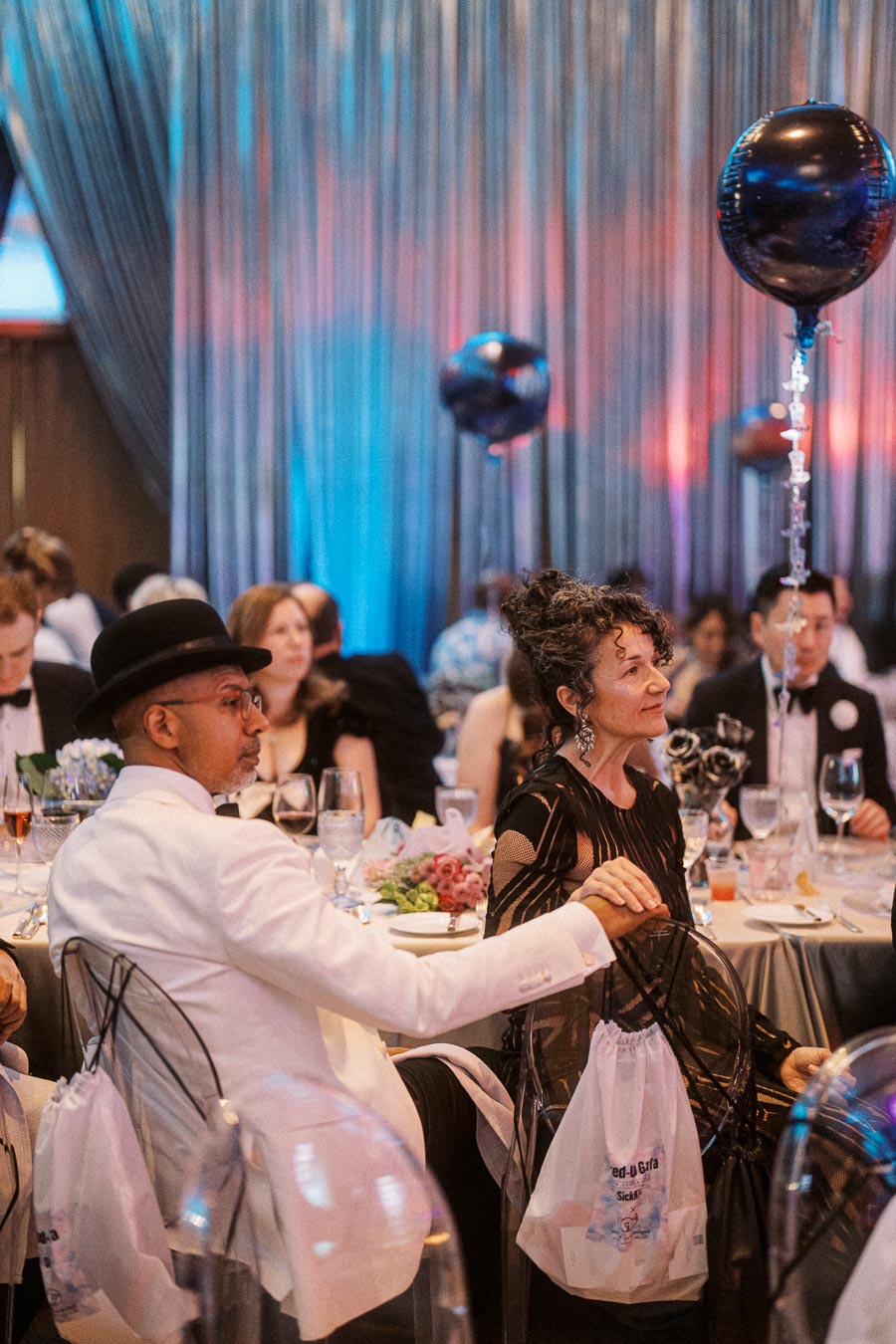 Elegant evening event with attendees in formal attire, seated at tables decorated with flowers and balloons, under ambient lighting.