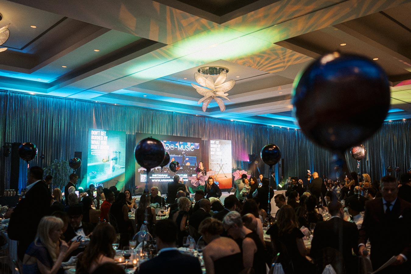 Elegant ballroom event with a glamorous crowd, colorful lighting, and stage with projections in the background. Balloons and an engaged audience create a lively atmosphere at the indoor celebration.
