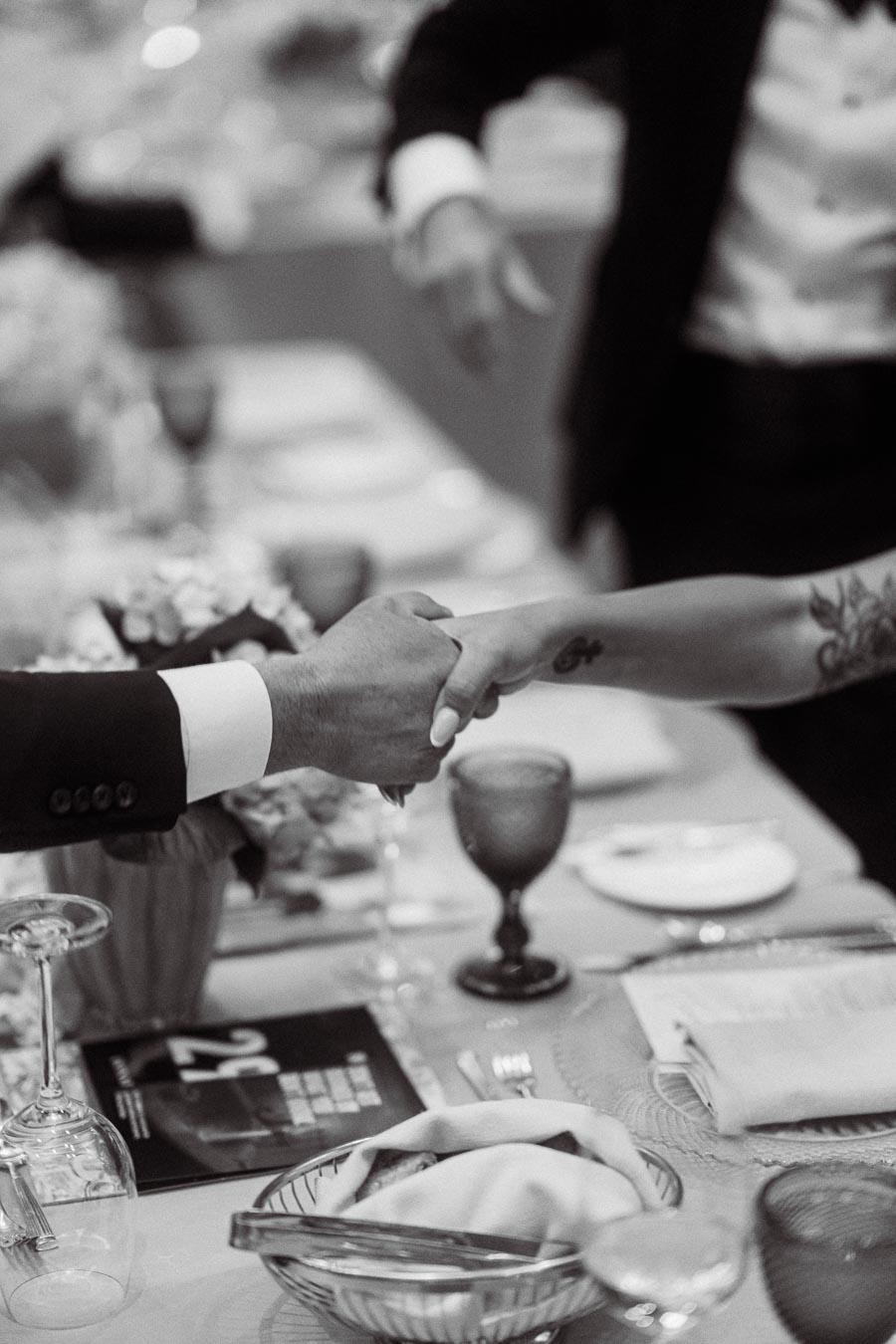 Black and white image of two people shaking hands over an elegantly set dinner table with wine glasses and flowers.