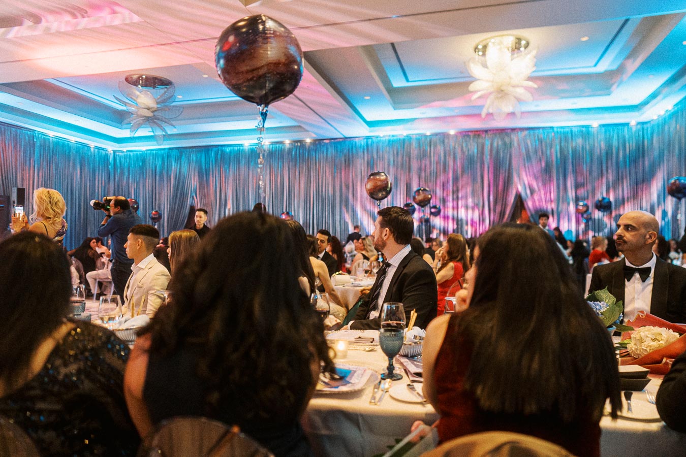 Elegant evening gala with guests seated at round tables under vibrant lighting, featuring unique balloon decorations and formal attire, creating a sophisticated event atmosphere.